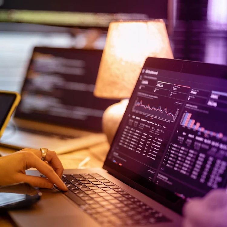 Profile of a woman working on various graphs on her computer. In the background, there is another blurred monitor.