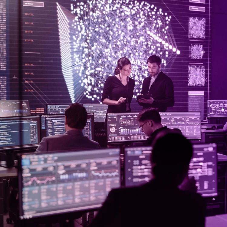Four professionals work in a high-tech control room. Two are standing and collaborating on a tablet in front of a large screen displaying a complex network of data and visualisations, while the other two are seated and monitoring information on different screens.